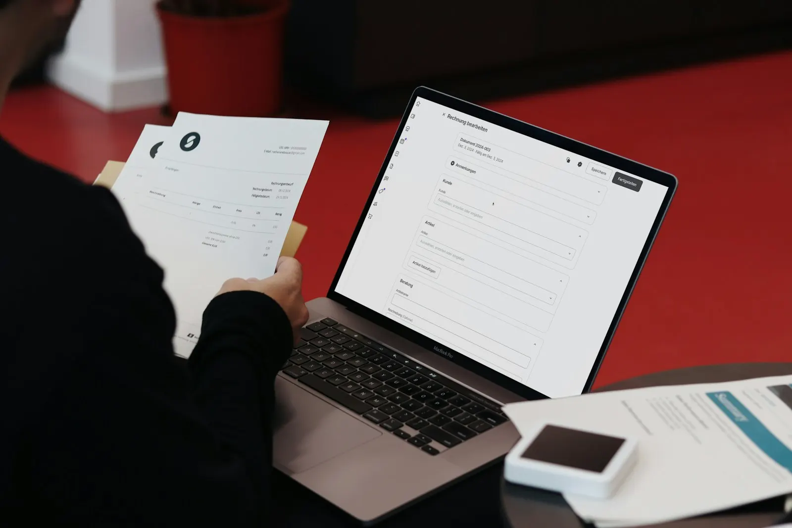 Person vid ett skrivbord som jämför pappersfakturor med ett fakturasystem på laptopen.